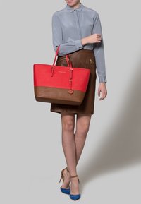 Červená a hnědá kabelka s koženou texturou, zlaté kovové akcenty a logo; v kombinaci se šedou blůzou a hnědými sukněmi. Viditelné modré podpatky.