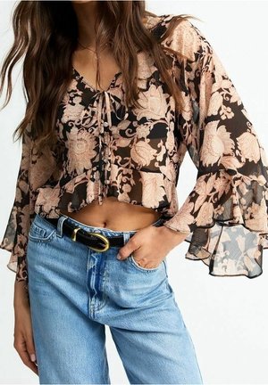 Blouse noire transparente à fleurs avec manches cloche et col à nouer, associée à un jean taille haute bleu clair et une ceinture noire avec boucle dorée.