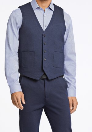 Man wearing navy blue buttoned vest with front pockets over light blue striped dress shirt and matching navy blue trousers.