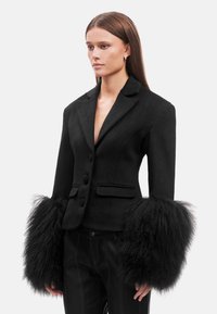 Blazer in lana nera con una silhouette aderente, caratterizzato da voluminosi dettagli in pelliccia nera sulle maniche e chiusura con bottoni. Dettagli di design su misura.