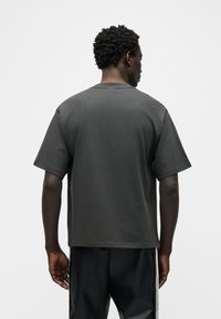 Νέος άνδρας με κοντά σγουρά μαλλιά που φοράει μια άνετη σκούρα γκρι μπλούζα και μαύρο παντελόνι με λευκές πλευρικές λωρίδες, γυρισμένος προς τα πίσω σε απλό φόντο.