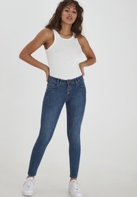 Jeans skinny bleus avec un devant à cinq boutons, des coutures contrastées et des détails subtils sur les poches ; associés à un débardeur blanc ajusté.