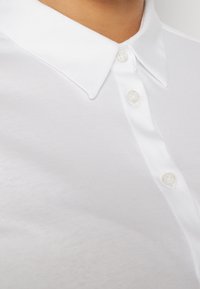 Gros plan sur une personne portant une chemise blanche avec col, boutonnée avec trois boutons blancs fermés.