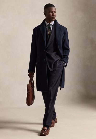 Homme en pardessus bleu marine, costume sombre, chemise rayée avec cravate à motifs, chaussures en cuir marron et portant une mallette en cuir marron marchant sur un fond neutre.