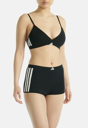 Frau trägt schwarzen Adidas-Sport-BH und passende Shorts mit weißen Streifen vor einem schlichten weißen Hintergrund.