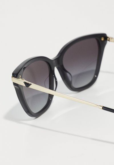 Ochelari de soare supradimensionați, negri, cu lentile gradient, rame din plastic groase și accente din metal de ton auriu pe tâmple.