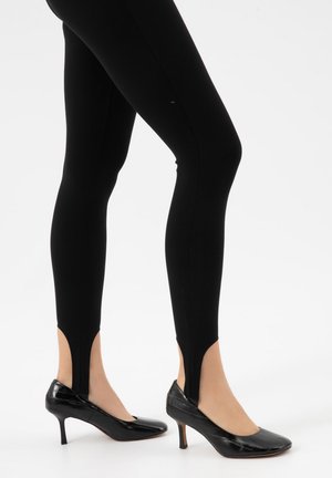 Benen in zwarte rijbroekleggings gecombineerd met zwarte lakleren pumps met een middenhoge hak op een witte achtergrond.