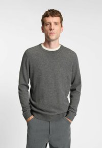 Maglione grigio lavorato a maglia con scollo rotondo e polsini a costine, abbinato a pantaloni grigi chiari. Il tessuto sembra morbido e strutturato.