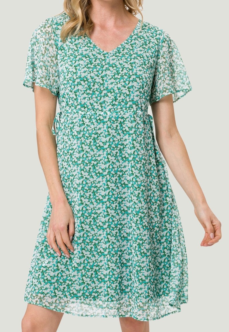 Vestido verde con escote en V y mangas cortas con volantes. Hecho de una tela ligera con un patrón de flores blancas y azul claro dispersas.