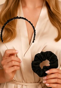 Czarna opaska na głowę z koralikami, z gładką metalową podstawą i czarną satynową gumką, trzymana przez osobę w beżowym szlafrok.