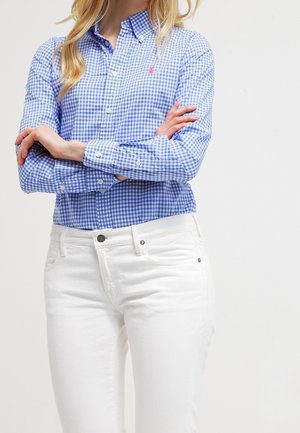 Femme portant une chemise à carreaux bleue et blanche à col et boutons, bras croisés, et jean blanc ajusté, sur un fond uni.