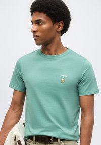 Νέος άνδρας φοράει ανοιχτό πράσινο T-shirt με μικρό έμβλημα ανανά και κείμενο στο αριστερό στήθος, κρατώντας ένα ανοιχτό σακάκι.