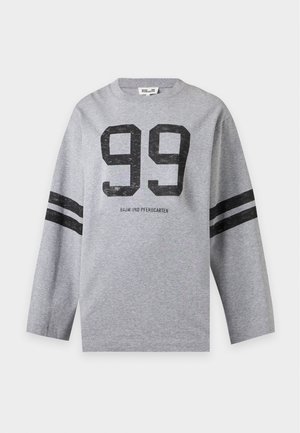 Femme portant un sweat gris avec le numéro 99 et des bandes noires sur les manches, associé à un pantalon blanc à jambes larges et des chaussures noires.