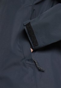 Detaliu al unei jachete waterproof de marină, arătând închiderea cu velcro la manșetă, accesul la buzunare și șnurul ajustabil, cu un model în carouri alb-negru.