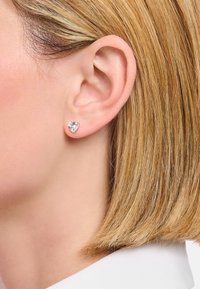 Herzförmige Steckerohrringe aus klarem Kristall, in einer Metallbasis in Silberoptik gefasst, getragen in einem kurzen Haarschnitt, der eine Nahaufnahme des Ohrs zeigt.