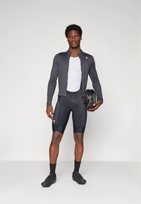 Pánsky cyklistický outfit pozostávajúci z čierneho dlhého trička s dlhým rukávom, bielej sieťoviny pod ním, čiernych šortiek a prilby. Navrhnutý pre pohodlie a výkon.