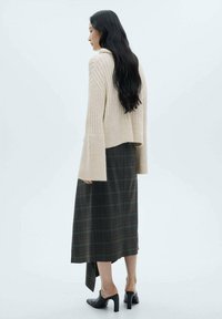 Maglione beige lavorato a maglia con maniche svasate, abbinato a una lunga gonna a quadri scura e sandali neri con tacco, visto da dietro.
