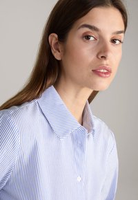 Blauw-wit gestreepte shirt met een puntige kraag, knoopsluiting en lange mouwen. De stof lijkt glad en lichtgewicht, met een subtiele glans.