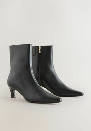 Bottines en cuir noir avec des orteils pointus, un talon fin et une fermeture éclair sur le côté avec un accent doré. Texture lisse et design épuré.
