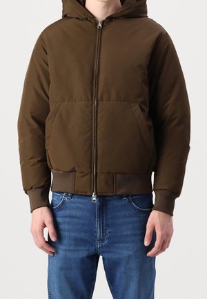 Homme portant une veste à capuche marron à fermeture éclair avec des poignets et un ourlet côtelés, associée à un jean en denim bleu, sur un fond uni.