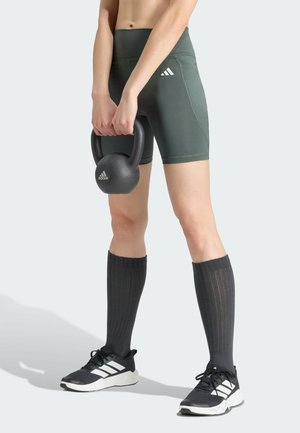 Persona che tiene un kettlebell nero con pantaloncini Adidas neri, calzini neri alti e sneakers Adidas nere su uno sfondo chiaro.