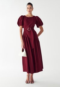 Bordeaux maxi jurk met pofmouwen, een vastgemaakte taille met een gouden gesp en een gerimpelde zoom. Gepaard met een witte handtas.