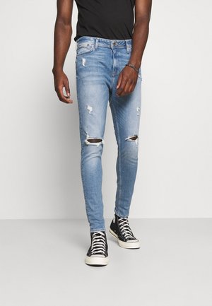 Homme portant un jean skinny bleu clair déchiré, des baskets montantes noires avec des lacets blancs et un t-shirt noir, debout devant un fond uni.