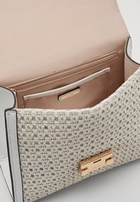 Tkaná kabelka v bílé barvě se zlatými kovovými prvky, s hladkým béžovým interiérem a kapsou na zip. Exteriér zdobí strukturovaný vzor.