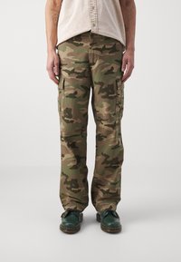 Camo cargo-broeken in groen- en bruintinten, gemaakt van stevig materiaal, met zijzakken en een losse pasvorm, gecombineerd met donkergrijze schoenen.