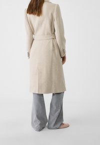 Vrouw met een beige mantel met ceintuur, grijze wijde jeans en roze schoenen, staand met de rug naar de camera tegen een effen witte achtergrond.