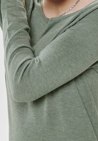 Haut vert à manches longues en tissu texturé doux, avec un col en V, des manches ajustées et un design minimaliste. Sans embellissements ni motifs visibles.