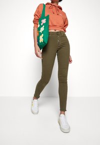 Henkilö kävelee oliivinvihreissä nappihousuissa, oranssissa lyhyessä hupparissa, valkoisissa tennareissa ja kantaa vihreää kankaislaukkua, jossa on värikkäitä kukkia.