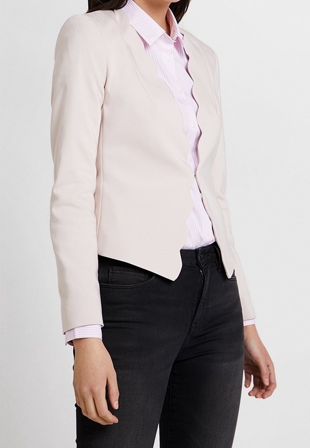 Blazer rose pâle sur mesure avec un col festonné, associé à une chemise rayée rose pâle à boutons et un jean noir, mettant en valeur une coupe ajustée.