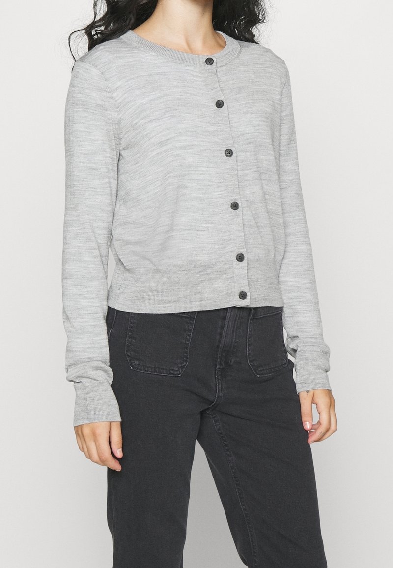 Cardigan tricoté gris clair avec des manches longues, un col rond et des boutons noirs sur le devant. Associé à un pantalon gris foncé.