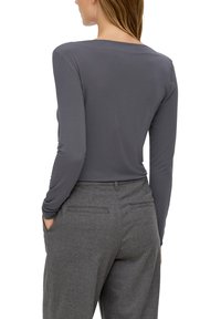 Vrouw in een nauwsluitend grijs shirt met lange mouwen en hoge taille grijze broek, met handen in de zakken, gezien van achteren.