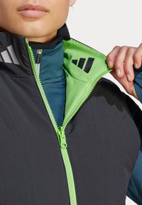 Zwarte sportieve vest met groene rits en binnenzijde. Bevat verticale lijnen en zwarte logo-accenten. Gelaagd over een teal shirt.