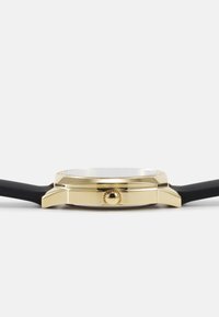 Gold-tone watch with a thin round case, featuring a black silicone strap, a textured crown, and a smooth glass face.