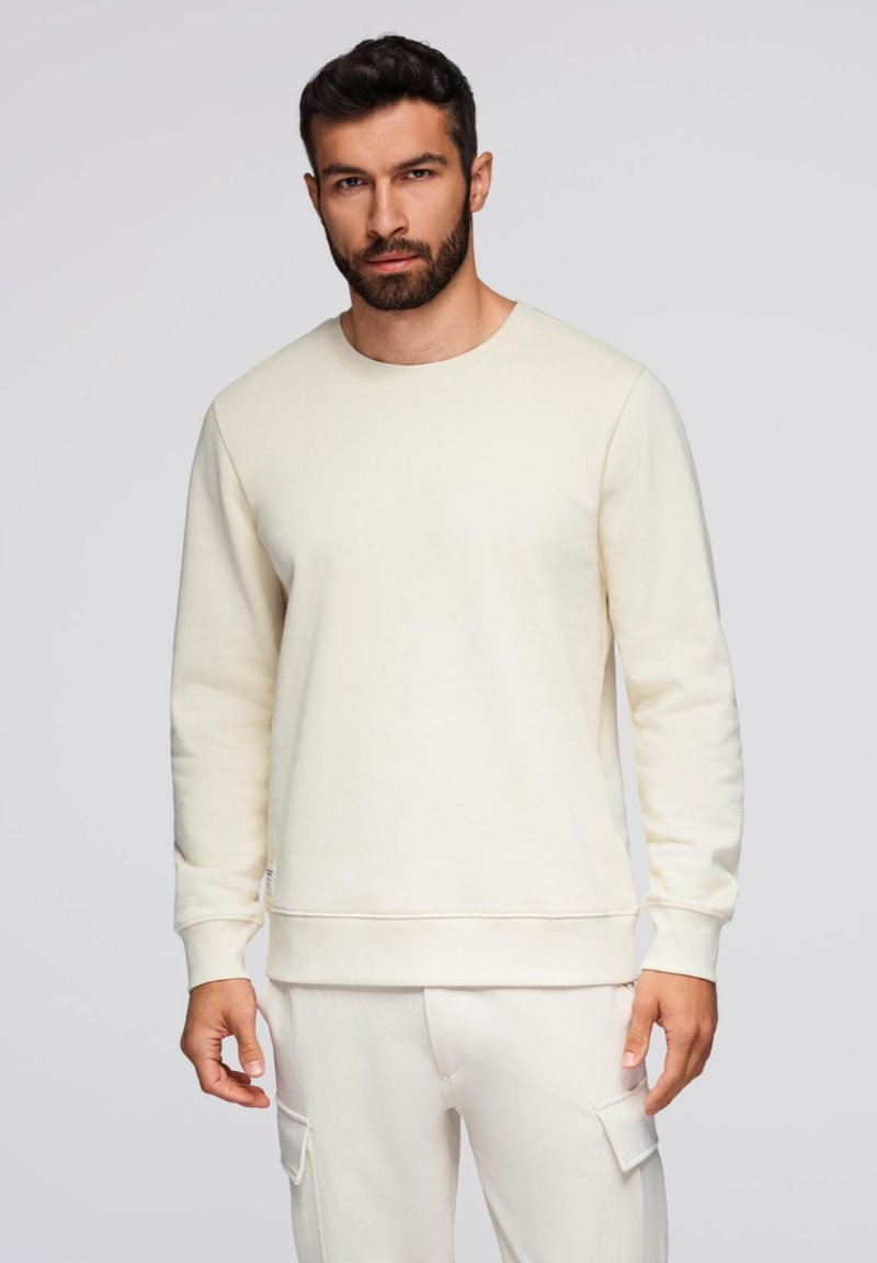 Kræmfarvet sweatshirt lavet af blødt stof, med rund halsudskæring og lange ærmer, samt ribbet talje og manchetter.