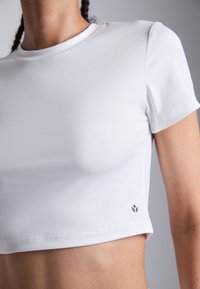 Hvit kort t-skjorte laget av glatt, elastisk materiale. Har korte ermer og en liten logo på nedre venstre side.