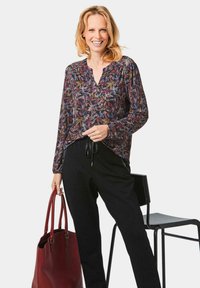 Langärmlige Bluse mit V-Ausschnitt und einem bunten Blättermuster auf dunklem Hintergrund, kombiniert mit schwarzen Hosen und einer roten Tragetasche.