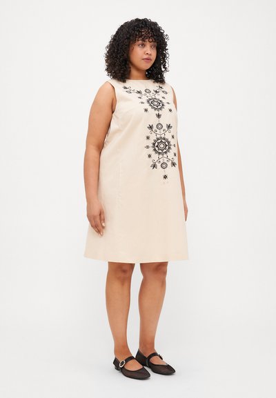 Robe sans manches beige avec broderie florale noire. Forme évasée, texture lisse, longueur genoux. Assortie avec des ballerines noires à boucle.