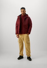 Maroon insulated jacket with a hood, layered over a red inner zip-up, paired with beige cargo pants featuring multiple pockets. Black footwear.