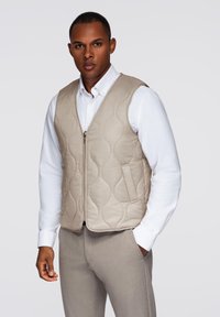Gilet trapuntato beige con chiusura a zip, caratterizzato da un design a scollo a V e texture liscia, indossato sopra una camicia bianca e pantaloni grigio chiaro.
