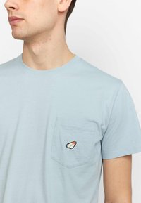 Lichtblauwe katoenen T-shirt met een ronde hals en korte mouwen, voorzien van een opgestikte pocket met een klein geborduurd ontwerp.