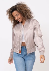 Bomberjack in licht beige satijn met geborduurde details, ritssluiting op de mouw en elastische manchetten. Draagt boven een witte top en blauwe jeans.