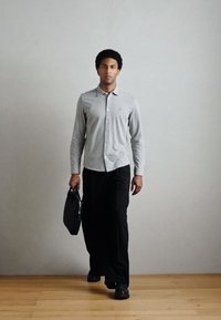 Camisa cinzenta de mangas compridas com botões, calças pretas e sapatos pretos. Segura uma pasta preta, de pé sobre um chão de madeira contra uma parede clara.