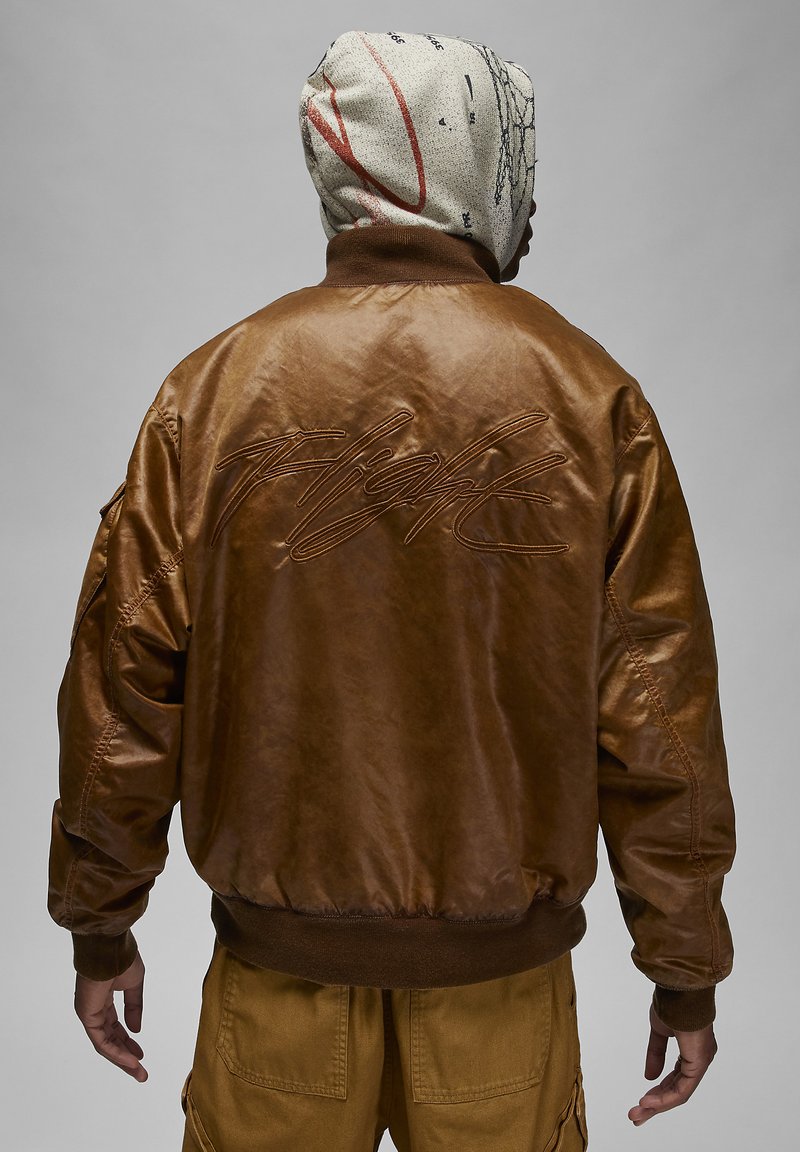 Braune Lederbomberjacke mit gerippten Bündchen, auf der Rückseite mit einem großen gestickten "Flight"-Design versehen. Getragen über khakifarbene Cargo-Hosen.