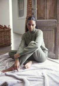 Zeleni pleteni pulover i usklađene hlače od mekanog materijala, sjedili na svijetlosivoj deki, s drvenim vratima i pletenom košarom u pozadini.