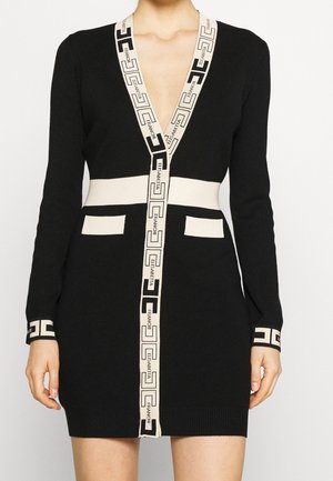 Zwarte gebreide jurk met lange mouwen, beige geometrische bies, voorzakken, diepe V-hals en taillebanddetail, gedragen door een staande persoon.