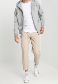 Homme marchant portant un sweat à capuche gris zippé, un t-shirt blanc, un pantalon beige retroussé et des baskets blanches à lacets sur un fond uni.